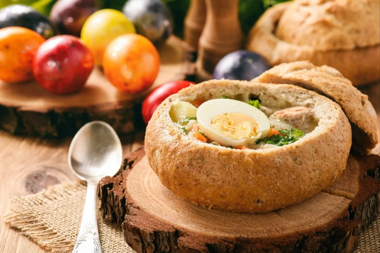 Tradycyjny żurek na zakwasie czy barszcz biały? Zupa podana w chlebie, z jajkiem i kiełbasą, obok kolorowe pisanki.