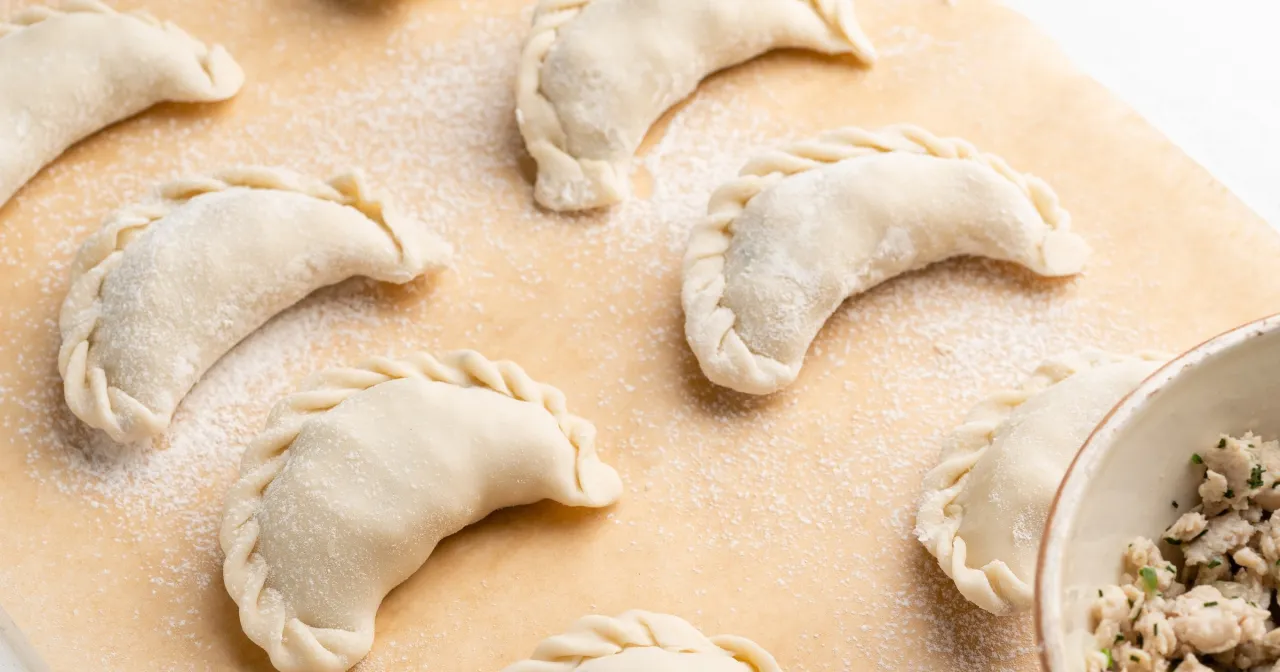 Surowe pierogi leżą na stolnicy, gotowe do wrzucenia do gotującej wody. Obok miseczka z farszem.