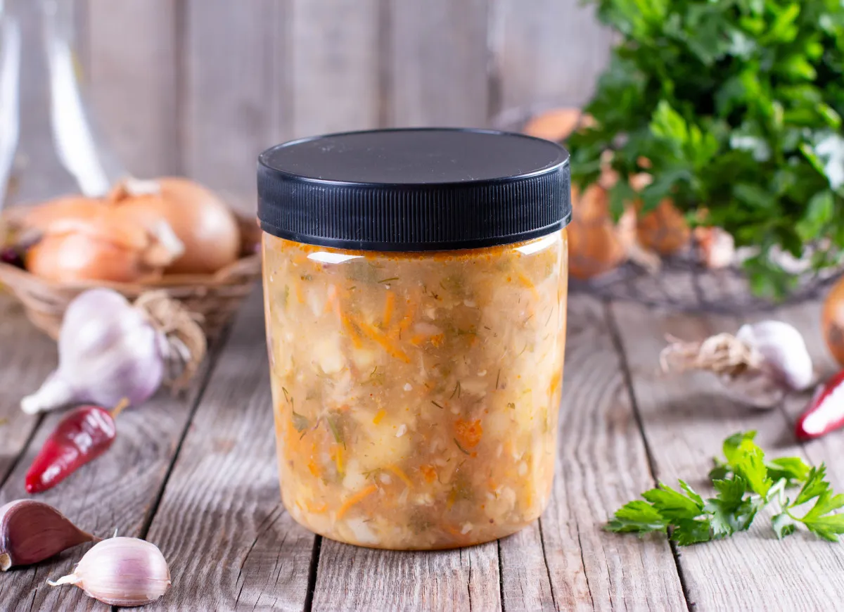 Słoik z zupą warzywną, obok cebula, czosnek i papryczka chili. Dowiedz się, jakich zup nie można mrozić, by uniknąć rozczarowania.