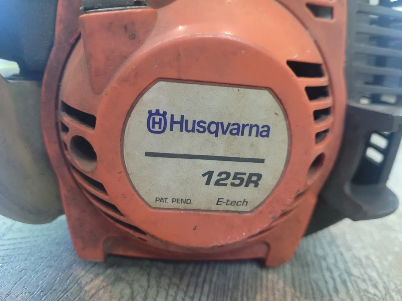 Pomarańczowa kosa spalinowa Husqvarna 125R z niebieskim logo. Gotowa do pracy w ogrodzie.