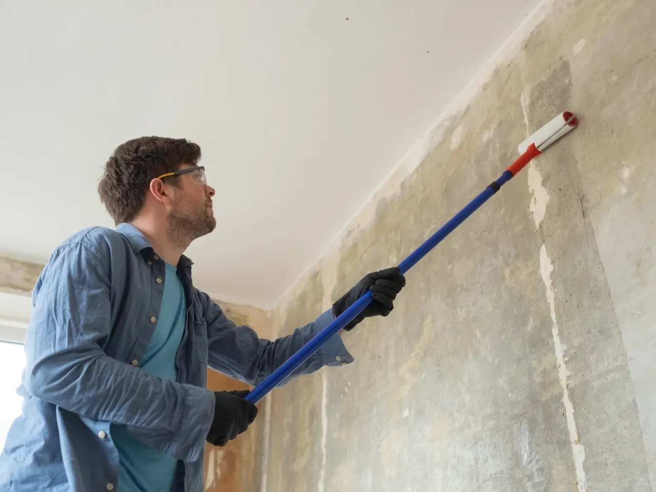 Mężczyzna w okularach ochronnych i rękawiczkach maluje ścianę wałkiem. Zastanawia się, ile razy malować ściany, by uzyskać idealny efekt.