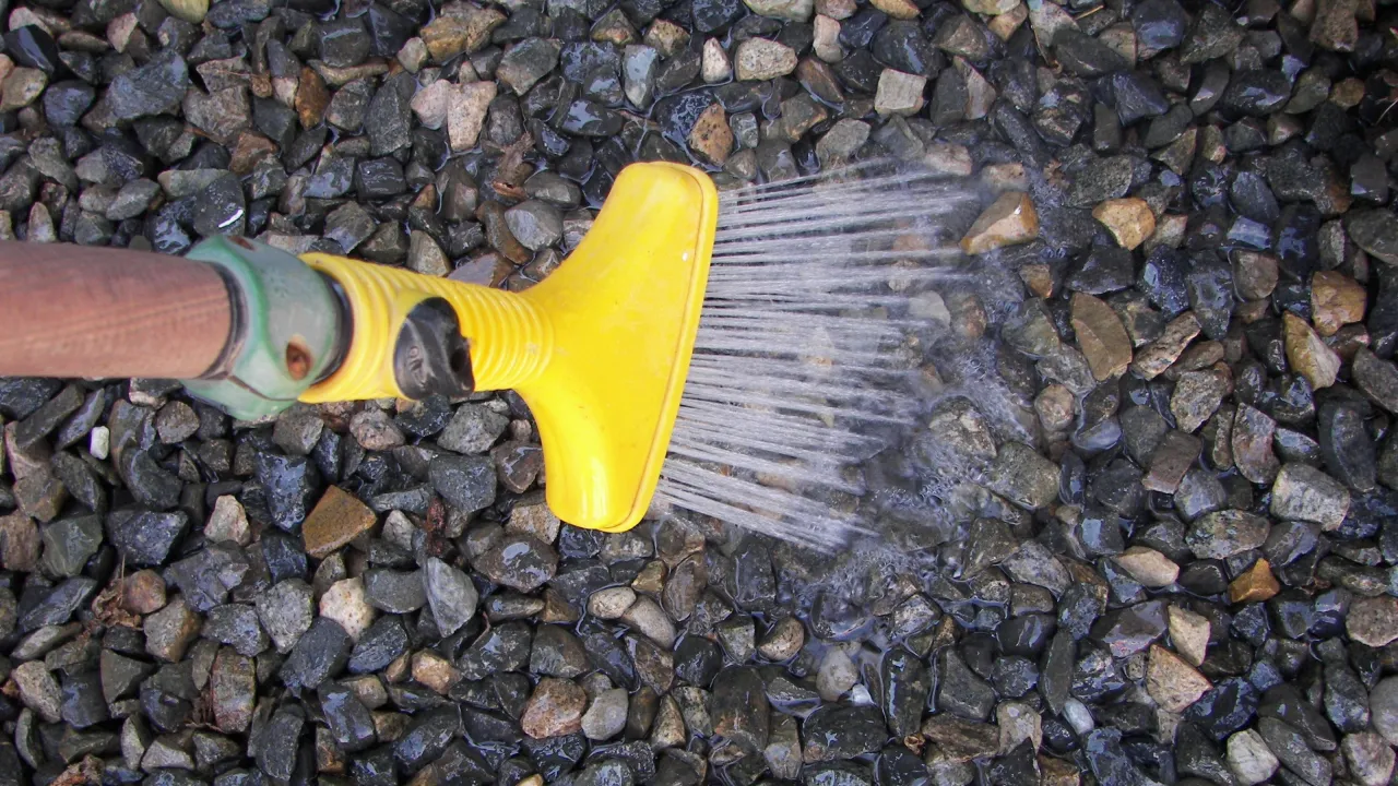 Ż&oacute;łta końc&oacute;wka węża ogrodowego spryskuje wodą kamyczki, pokazując, jak wyczyścić kamyczki w ogrodzie.