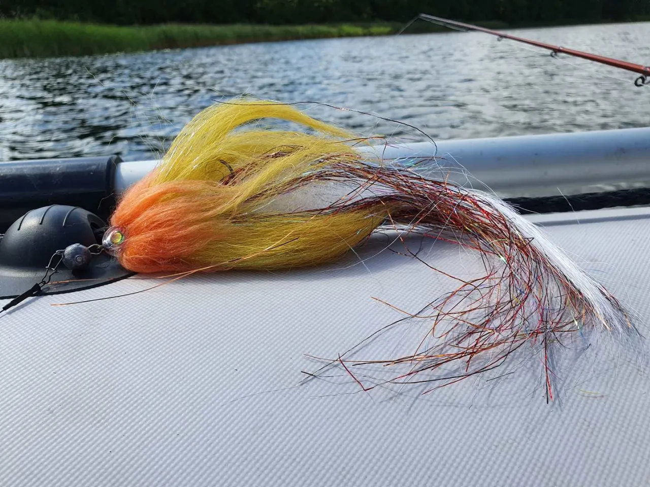Kolorowa mucha, idealna zanęta na szczupaka, leży na pontonie. W tle widać wodę i wędkę.