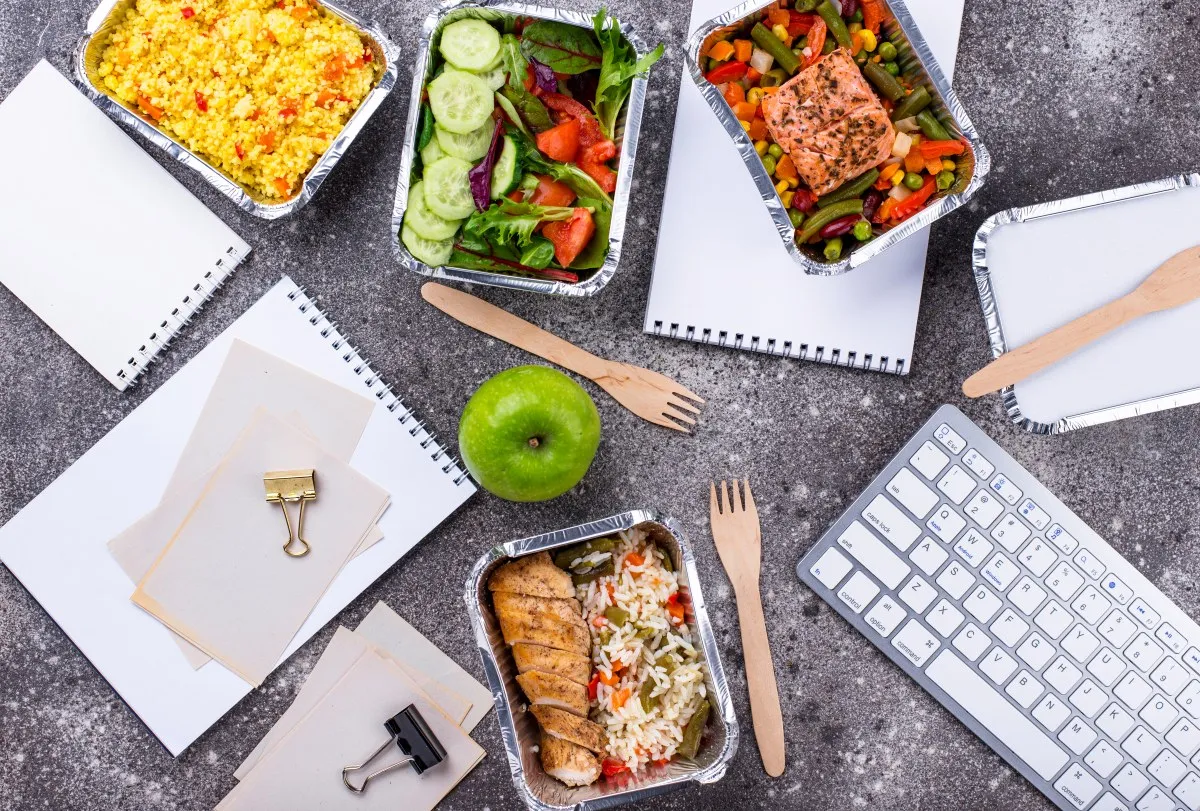 Zdrowy lunch vs obiad w pudełkach: kurczak z ryżem, łosoś z warzywami, sałatka, kasza. Obok jabłko, notatniki i klawiatura.