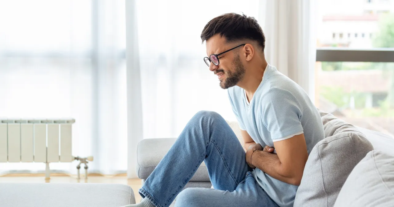 Mężczyzna w okularach siedzi na kanapie, trzymając się za brzuch. Wygląda na to, że szuka czegoś na b&oacute;l brzucha.