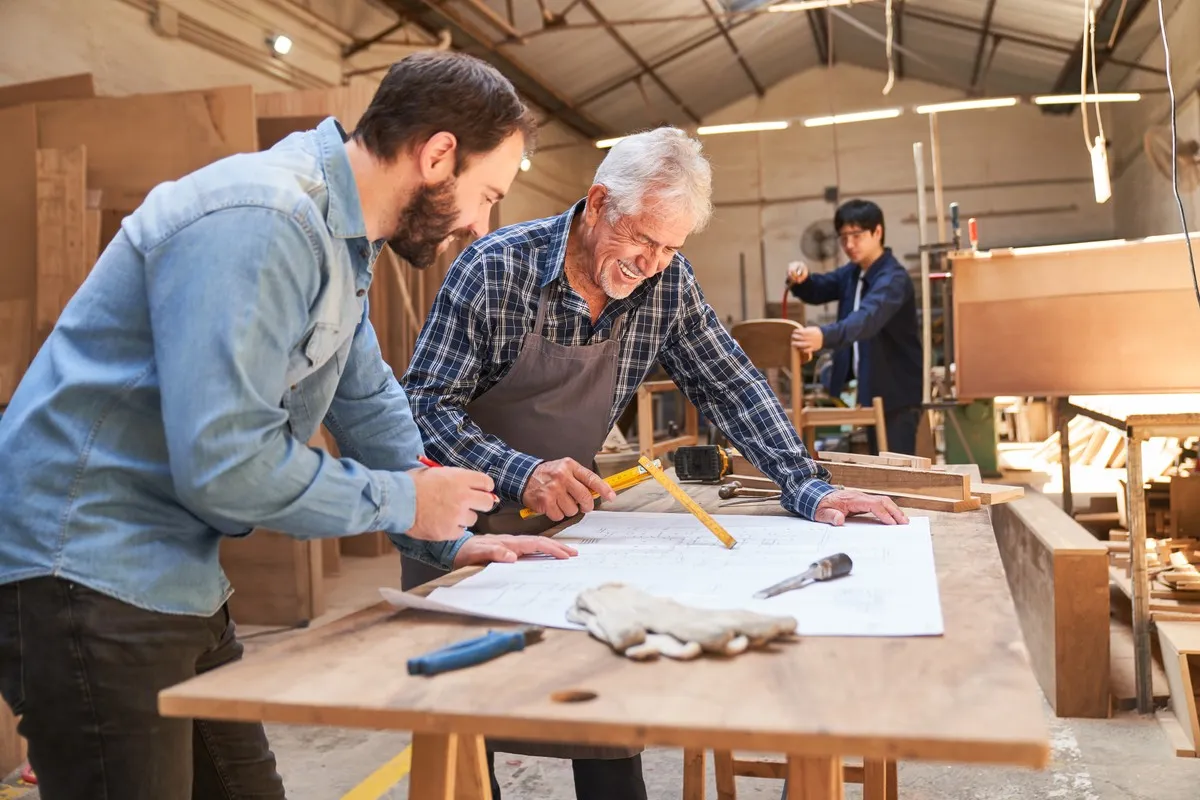 Uśmiechnięty senior z siwymi włosami, w kratę i fartuchu, z młodszym mężczyzną analizuje plany w stolarni.