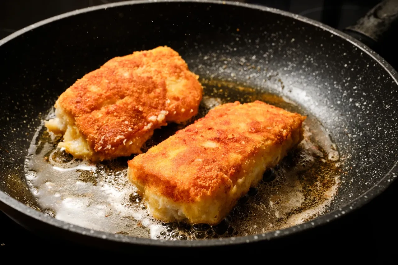 Dwa kawałki ryby w złocistej panierce smażą się na głębokim oleju na czarnej patelni. Idealne na czym smażyć rybę w panierce.