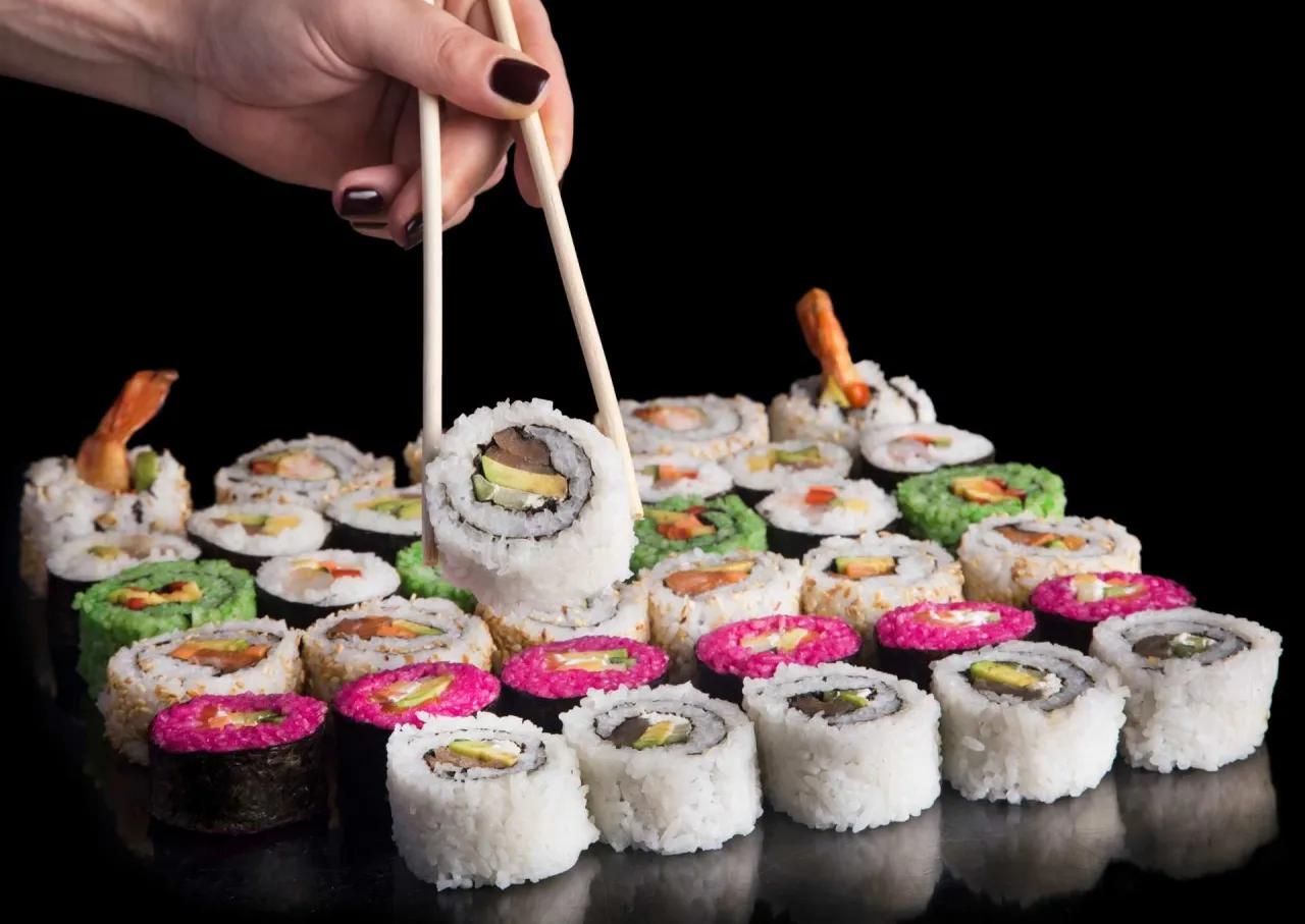 Dłoń z pałeczkami do sushi podnosi jeden z roladek. Wok&oacute;ł mn&oacute;stwo kolorowych sushi.