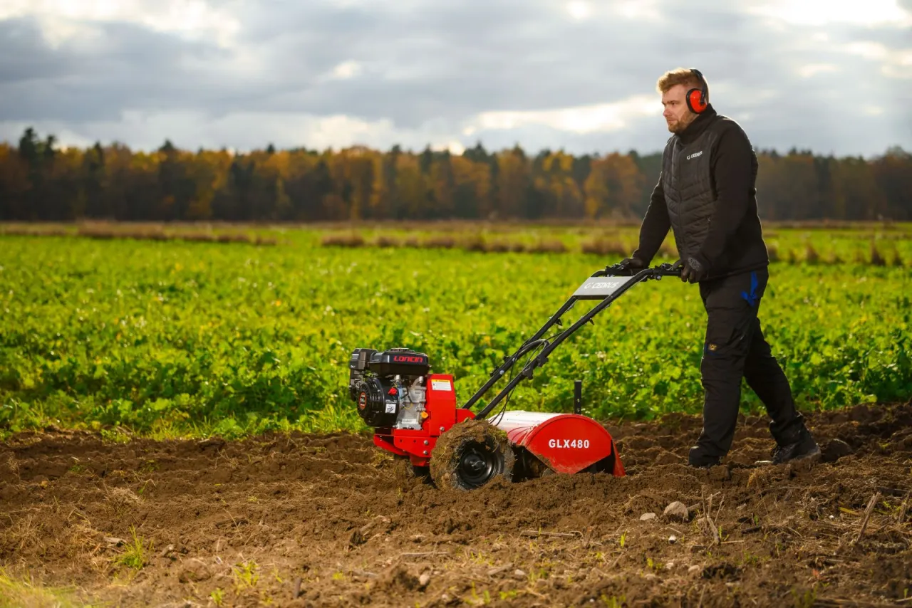 Mężczyzna w słuchawkach ochronnych pracuje glebogryzarką na polu. Pokazuje, jak używać glebogryzarki do spulchniania ziemi.