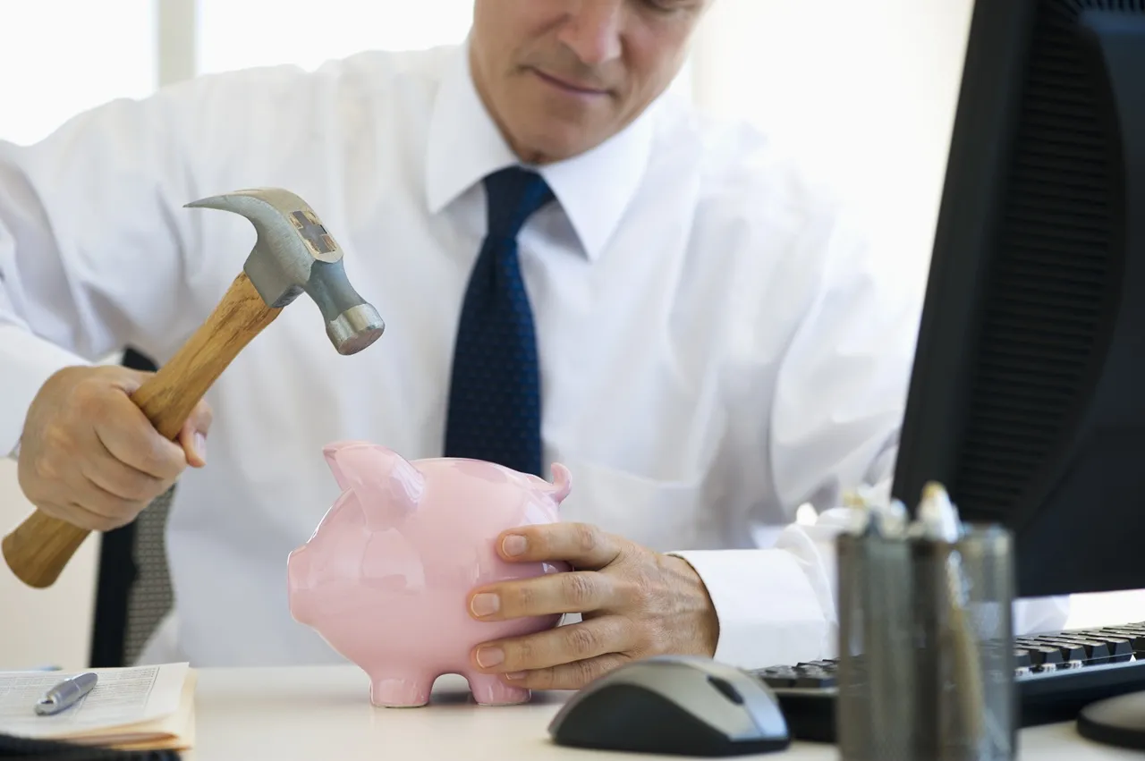 Mężczyzna w białej koszuli i krawacie zamierza rozbić skarbonkę-świnkę młotkiem. Zastanawia się, ile trwa likwidacja lokaty, gdy pieniądze są tak trudno dostępne.