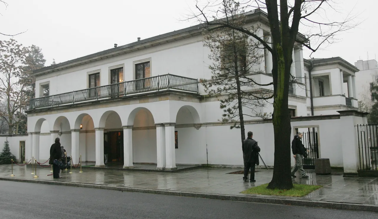 Biała willa z arkadami i balkonem, gdzie może mieszkać Tusk. Na zewnątrz kilka osób, w tym dziennikarz z kamerą.