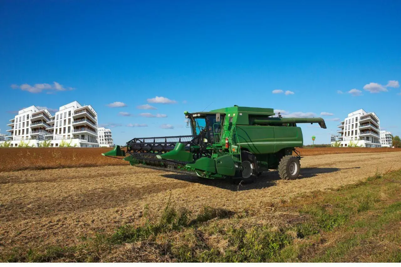 Zielona kombajn na polu, w tle nowoczesne budynki. Obraz symbolizuje zmieniającą się rzeczywistość i liczbę rolnik&oacute;w w Polsce.