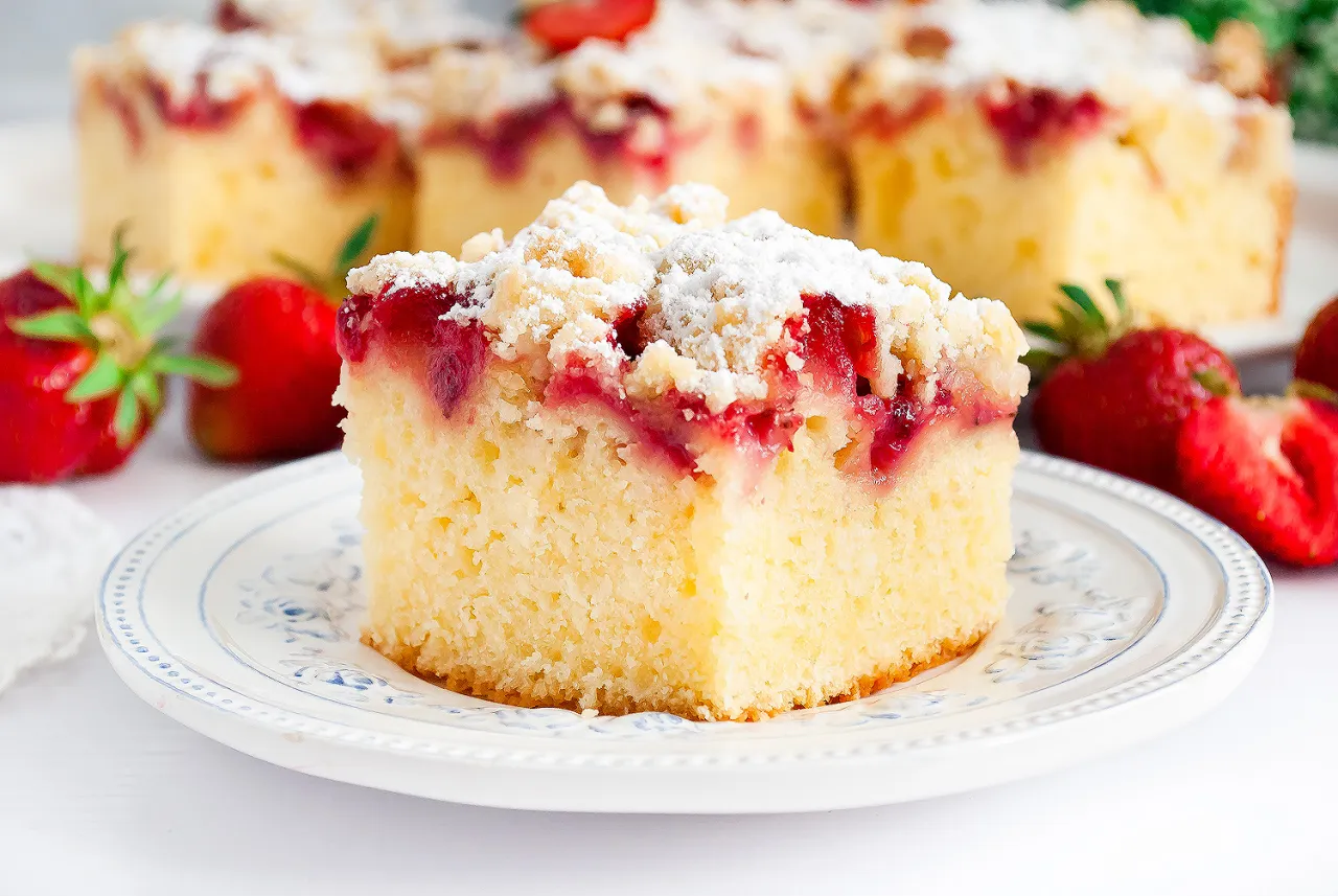 Puszyste ciasto jogurtowe z truskawkami, posypane cukrem pudrem i kruszonką. Obok świeże truskawki.