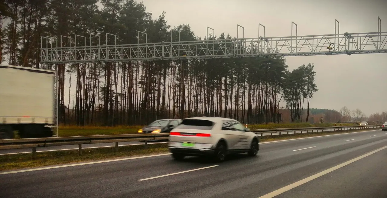 Na drodze S11 widać bramownicę do odcinkowego pomiaru prędkości. Samochody jadą w kierunku lasu.