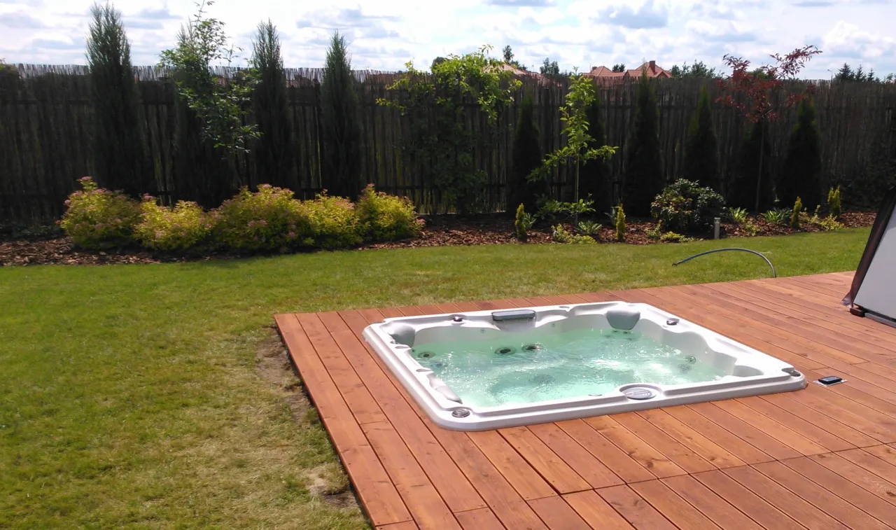 Jak zabudować jacuzzi ogrodowe? Biała wanna z hydromasażem na drewnianym tarasie, otoczona zielenią i płotem z bambusa.