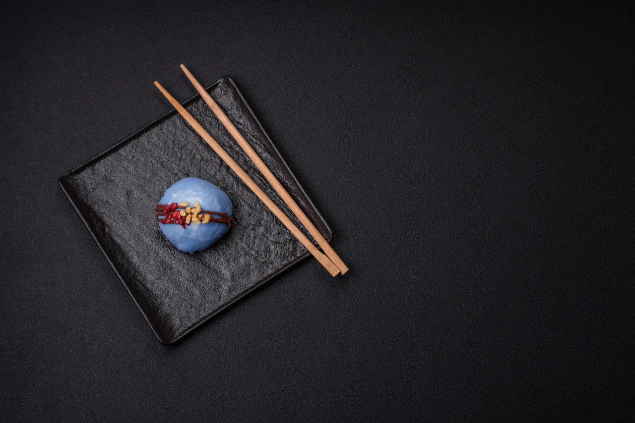 Niebieska kulka z polewą czekoladową i posypką na czarnej zastawie, obok pałeczki. Piękne sushi kompozycje.