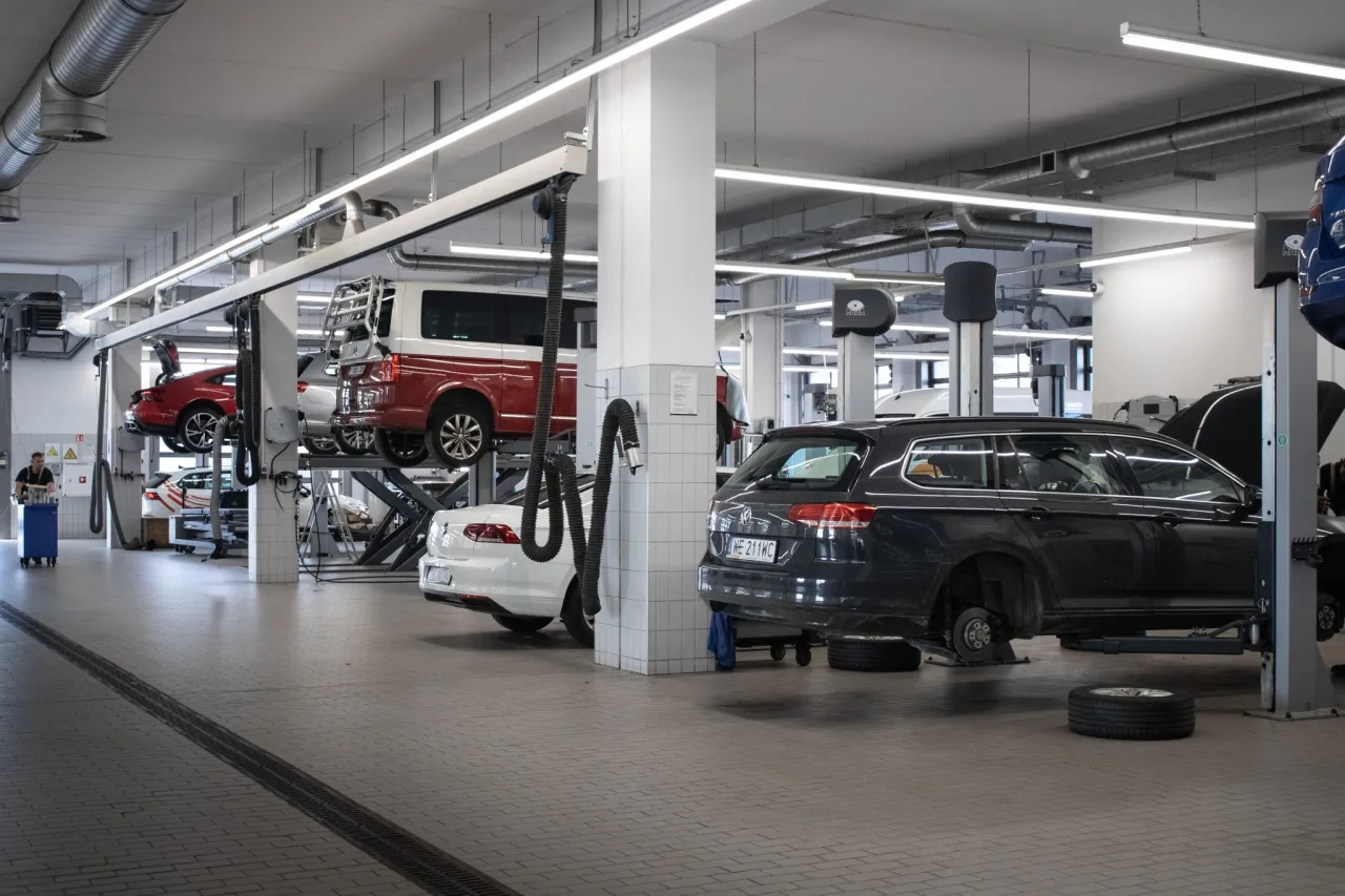 Warsztat samochodowy z kilkoma autami na podnośnikach. Widać czerwony van, białe i ciemne kombi. Mechanik pracuje przy panelu sterowania. To dobry aso skr&oacute;t.