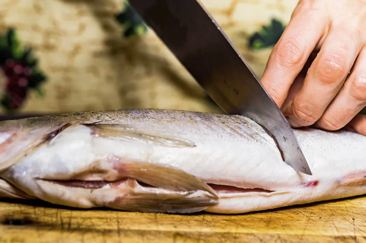Przygotowanie karpia do zamrożenia: ostry n&oacute;ż kroi rybę na desce.
