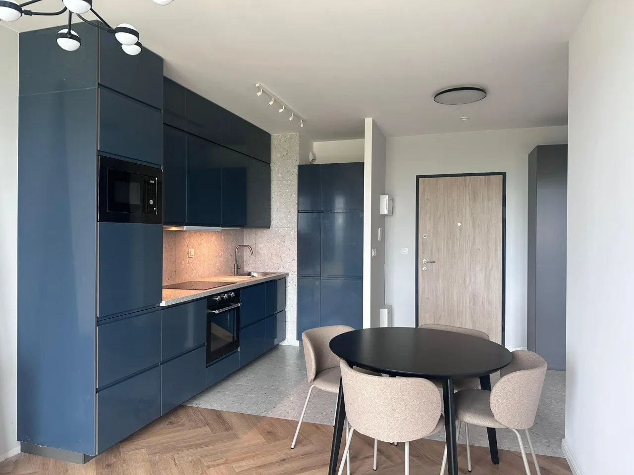 Nowoczesne wykończenie mieszkań Warszawa: granatowa kuchnia z wyspą, jadalnia z okrągłym stołem i krzesłami, drewniana podłoga.