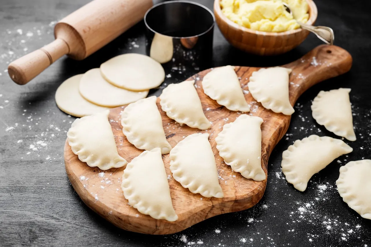 Gotowe do lepienia pierogi na desce, obok wałek, wykrawaczka i miska z farszem. Idealny przepis na to, jak lepić pierogi.