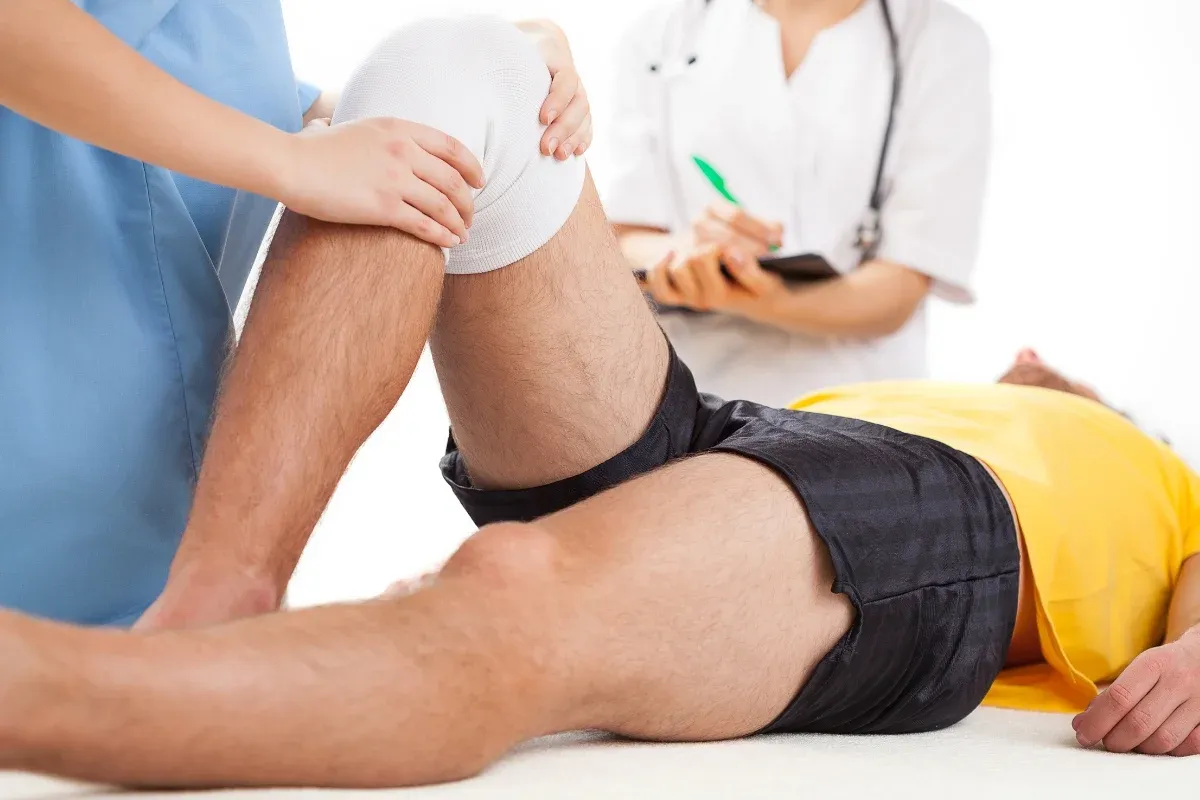 Fizjoterapia kolana na czym polega? Terapeuta delikatnie uciska opatrunek na kolanie pacjenta, podczas gdy lekarz notuje postępy.