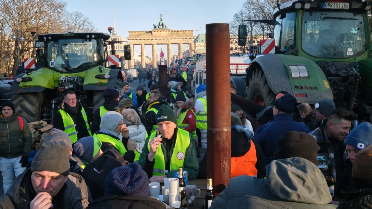 Rolnicy protestują pod Bramą Brandenburską, pytając, co to jest zielony ład w rolnictwie. W tle widać traktory i tłum ludzi.