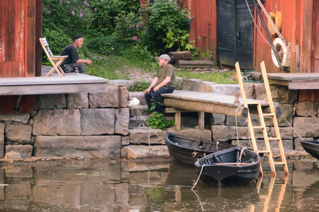 Dw&oacute;ch mężczyzn odpoczywa nad wodą, w tle drewniana chata i bujna zieleń. Scena jak z pojezierza fińskiego.