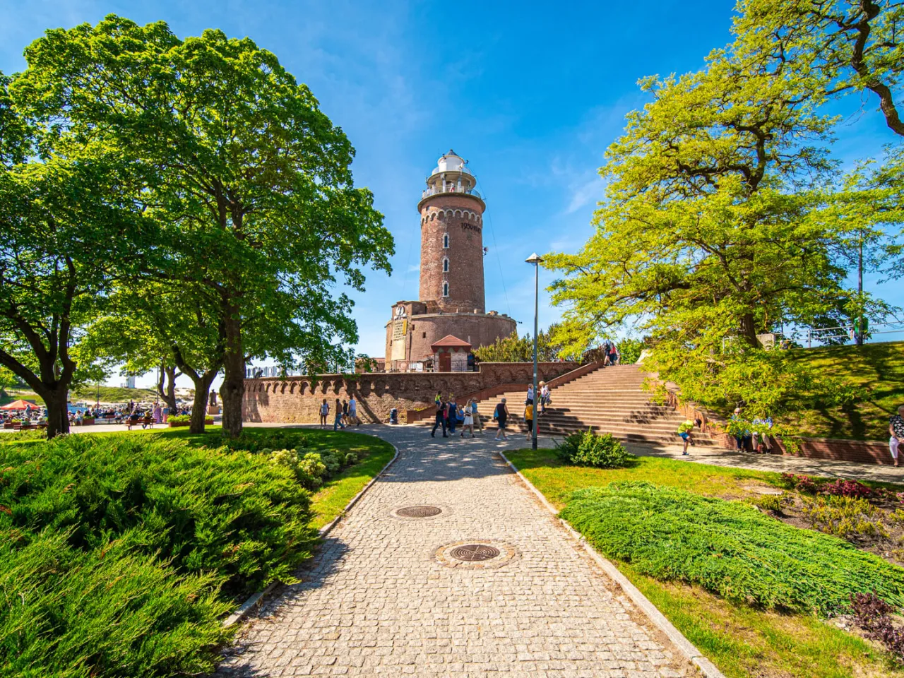 Latarnia morska w Kołobrzegu, jeden z punktów, co warto zobaczyć w Kołobrzegu, otoczona zielenią i kamienną ścieżką.