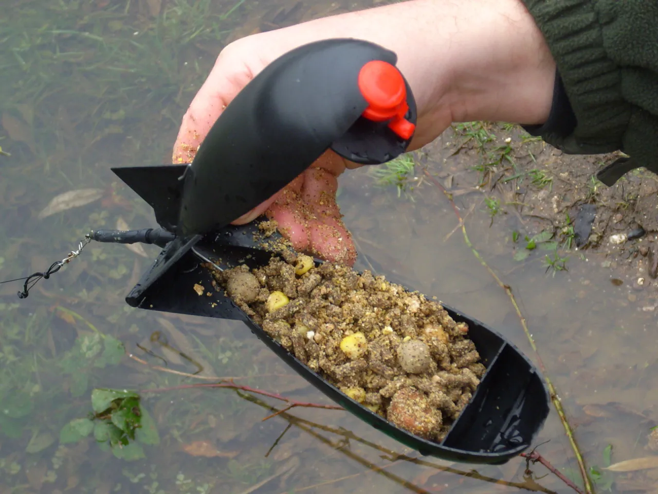 Ręka trzyma otwarta rakietę z zanętą do zig riga, gotową do zarzucenia do wody.