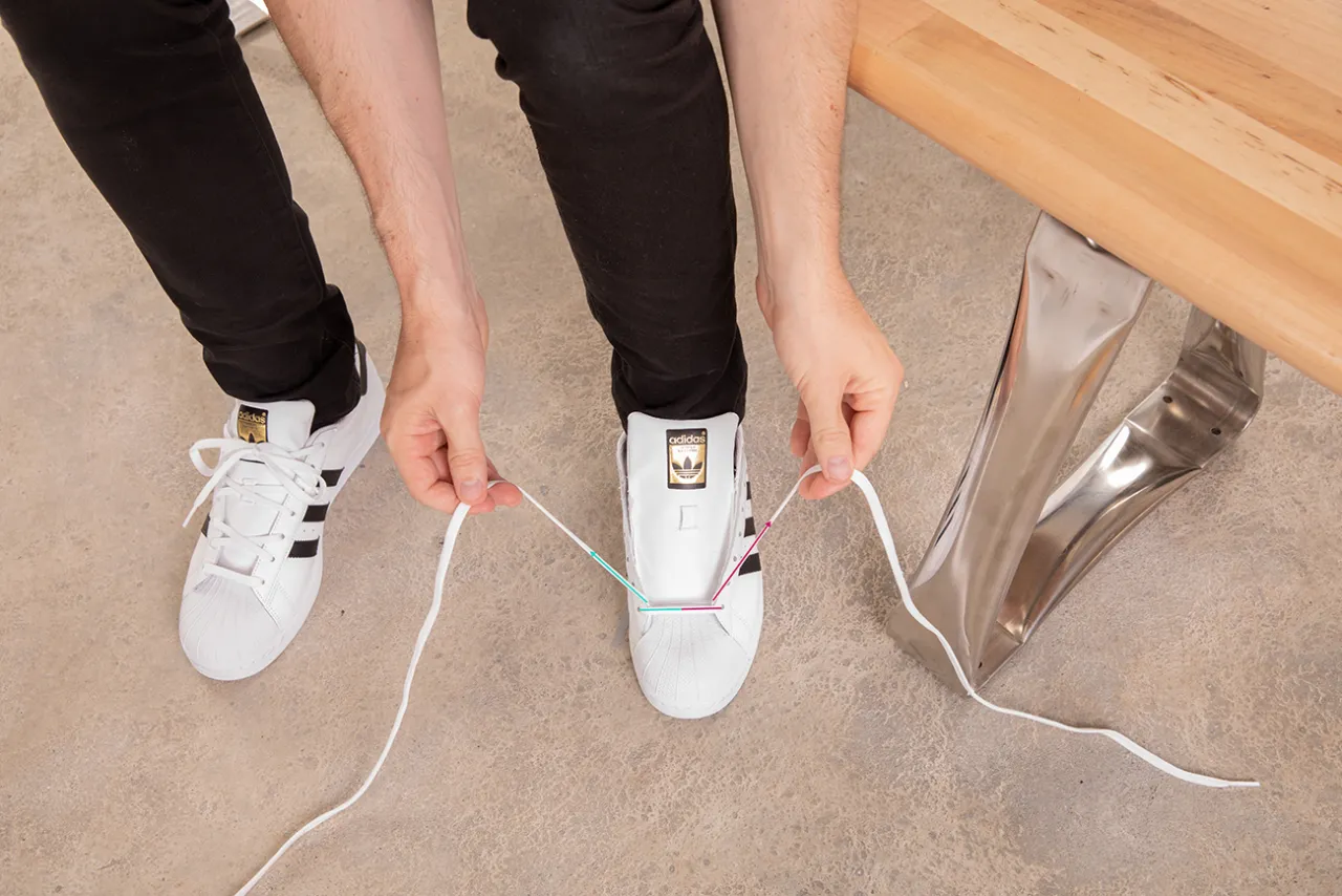 Instrukcja krok po kroku, jak wiązać sneakersy. Dłonie trzymają białe sznurówki, przygotowując się do zawiązania butów Adidas.