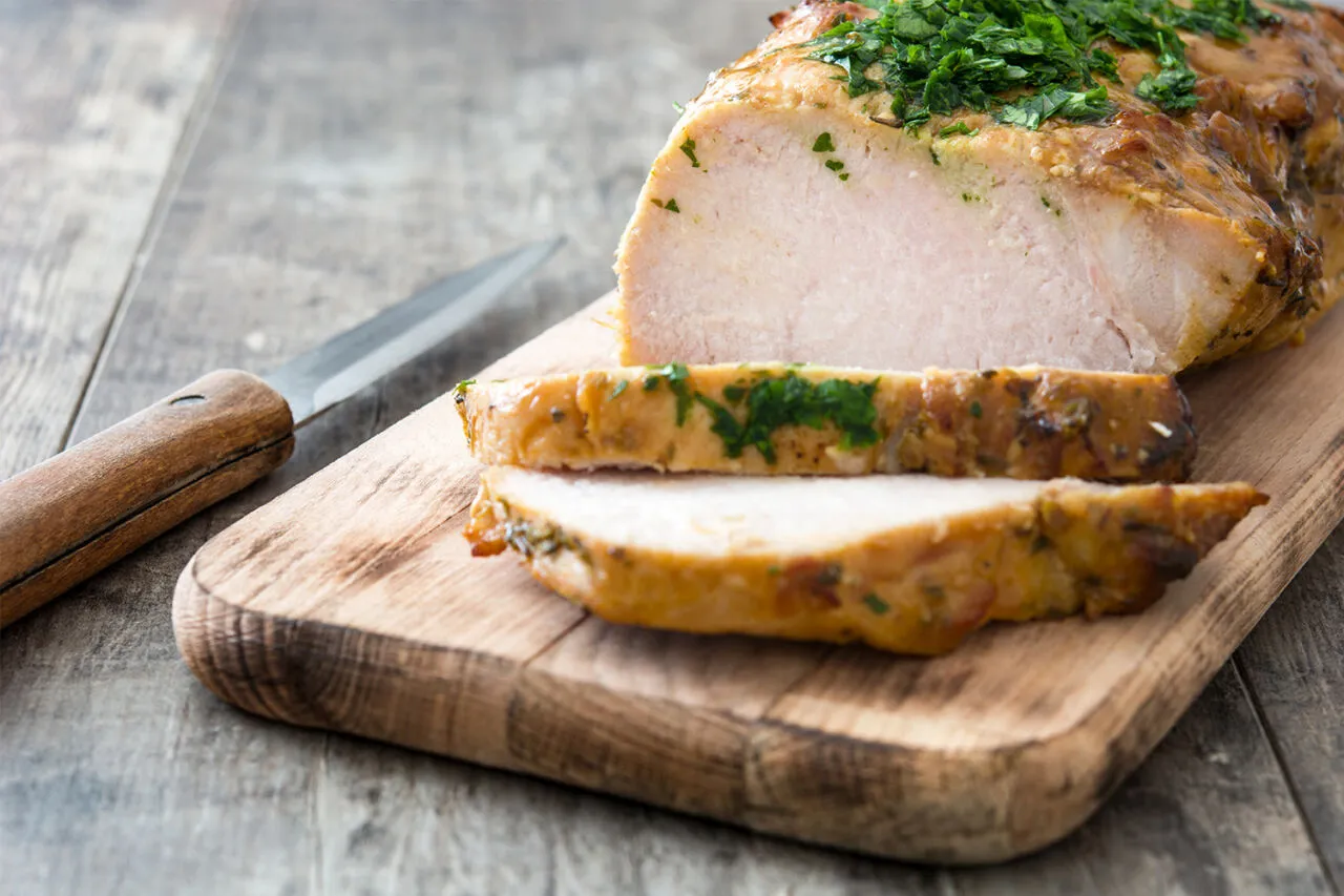 Pyszny, pieczony schab, pokrojony w plastry, z ziołami na wierzchu, leży na desce obok noża.