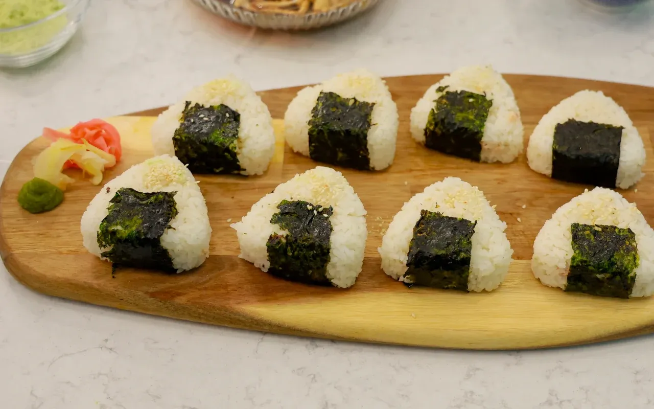 Na desce leży osiem tr&oacute;jkątnych onigiri z tuńczykiem, owiniętych w nori, posypanych sezamem. Obok znajduje się wasabi i marynowany imbir.