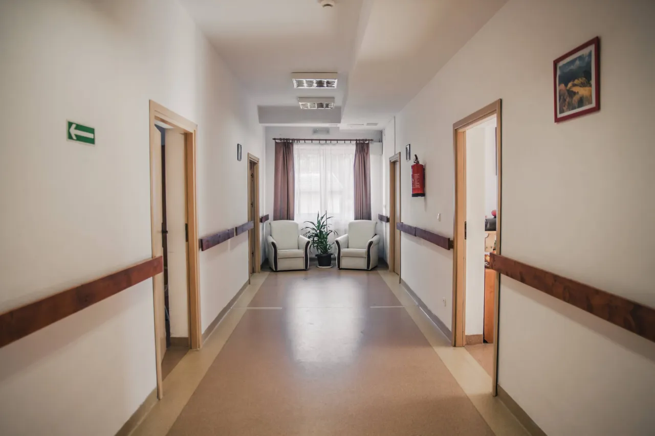 Jasny korytarz zakładu opiekuńczo-leczniczego z poręczami, fotelami i roślinami. Sprawdź wolne miejsca.