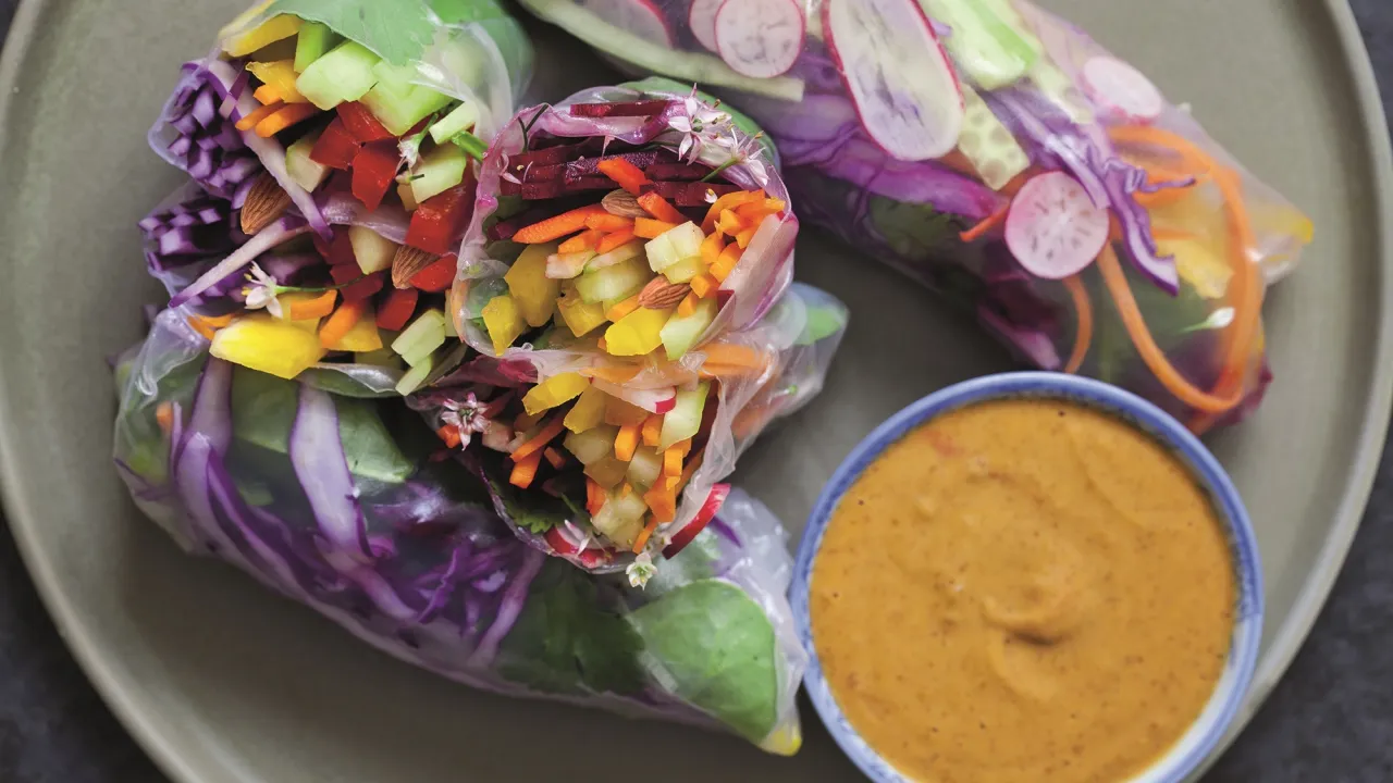 Kolorowe rolki z papieru ryżowego z warzywami: marchewka, og&oacute;rek, papryka, czerwona kapusta, rzodkiewka. Obok miseczka z sosem.