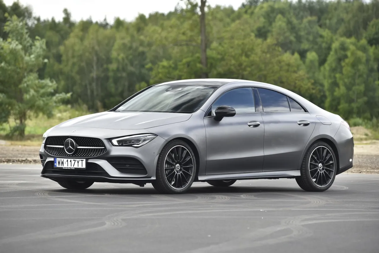 Elegancki, szary Mercedes-Benz CLA Coupe z czarnymi felgami stoi na drodze, z lasem w tle.
