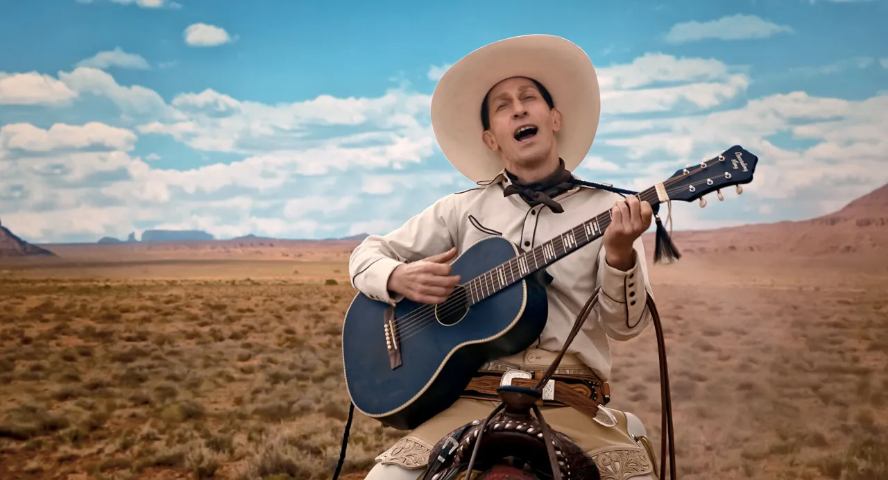 Tim Blake Nelson jako Buster Scruggs w westernowym stroju gra na gitarze na tle pustynnego krajobrazu. Ballada o Busterze Scruggsie obsada.
