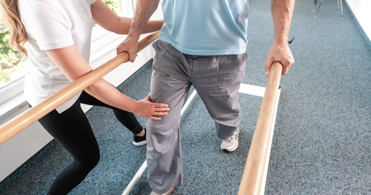 Fizjoterapeuta pomaga pacjentowi w rehabilitacji przy poręczach. Dowiedz się, jak obliczyć dofinansowanie do turnusu rehabilitacyjnego.
