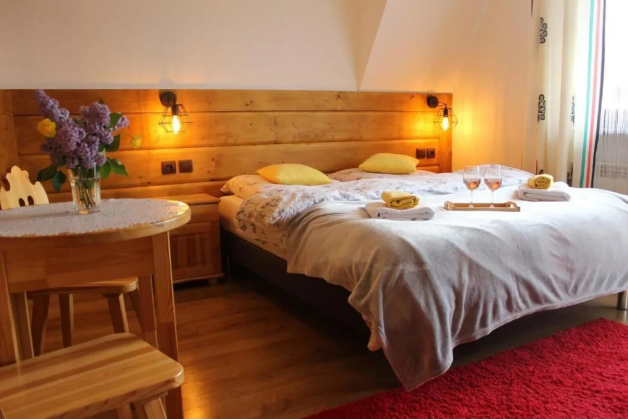 Przytulny pok&oacute;j w willi pod Zielonym Wierchem w Zakopanem. Ł&oacute;żko z ż&oacute;łtymi poduszkami, bukiet bzu i kieliszki z winem.