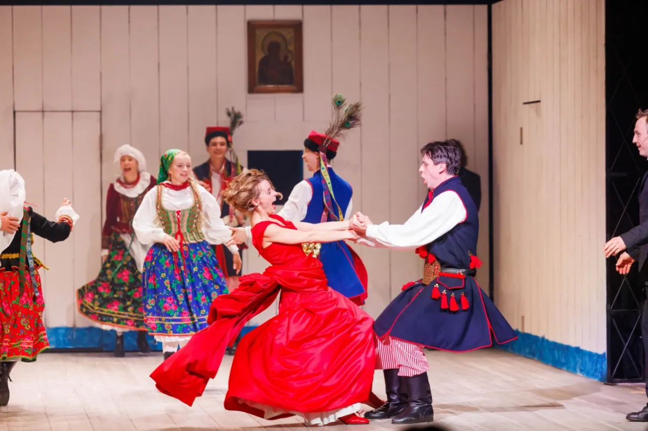 Tancerze w strojach ludowych, w tym kobieta w czerwonej sukni, tworzą żywiołową syntezę sztuk w weselu na scenie.
