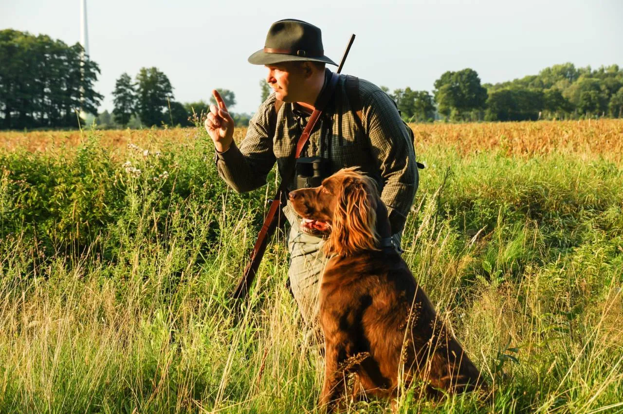 Myśliwy w kapeluszu i z psem w polu, daje znak.
