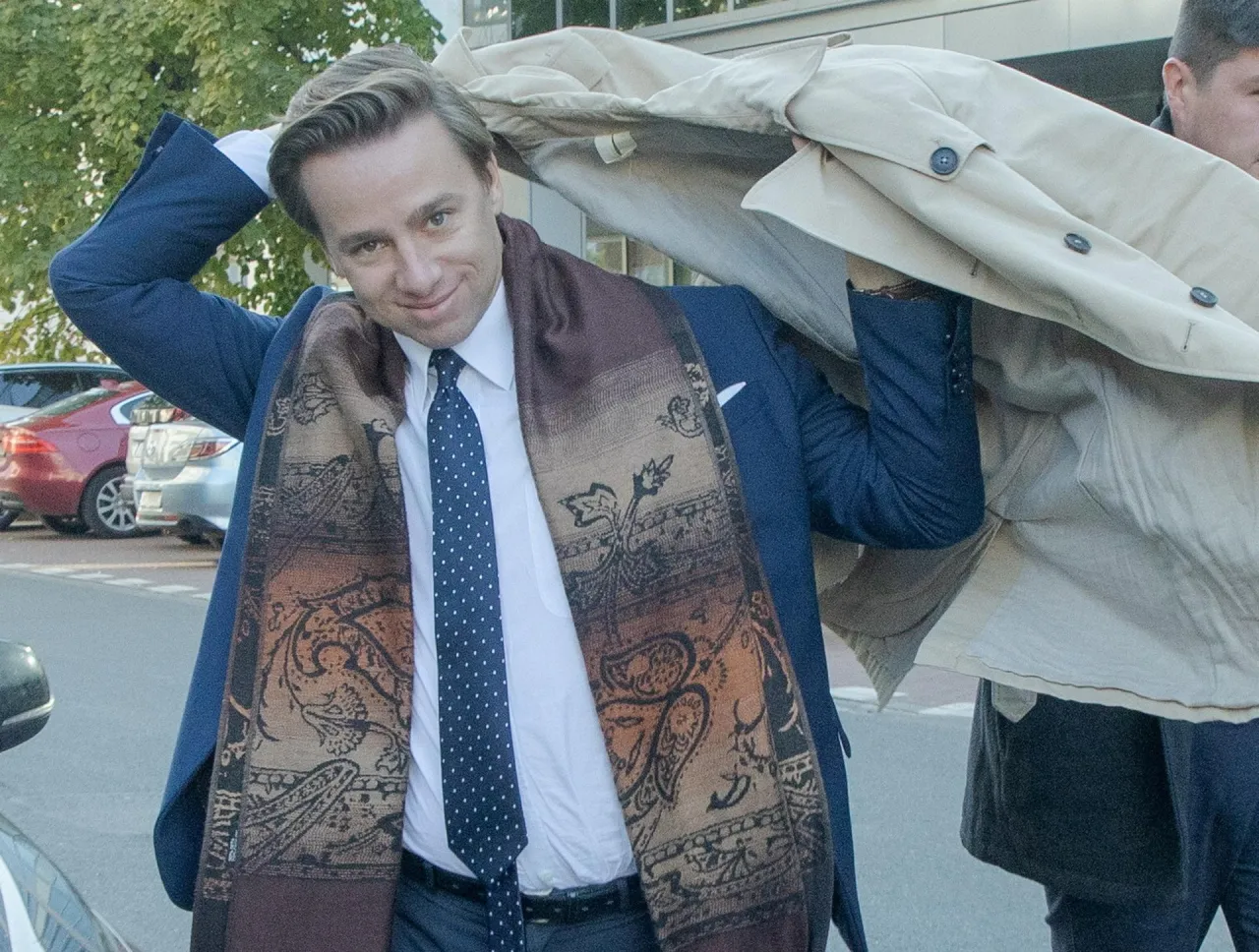 Mężczyzna w garniturze i szalu z wzorem, który mógłby mieć ile lat ma bosak, uśmiecha się, trzymając płaszcz.