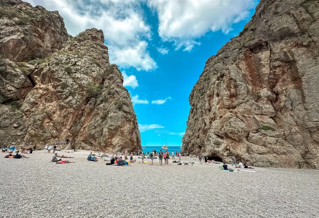 Plaża w wąwozie, otoczona skalnymi ścianami, to jedno z najpiękniejszych miejsc na Majorce. Ludzie odpoczywają na kamienistym brzegu, a w oddali widać łodzie.