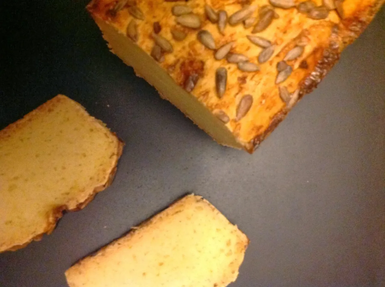 Kawałek chleba bezglutenowego z pestkami słonecznika na wierzchu i dwa plastry obok. Idealny przykład, jak upiec chleb bezglutenowy.