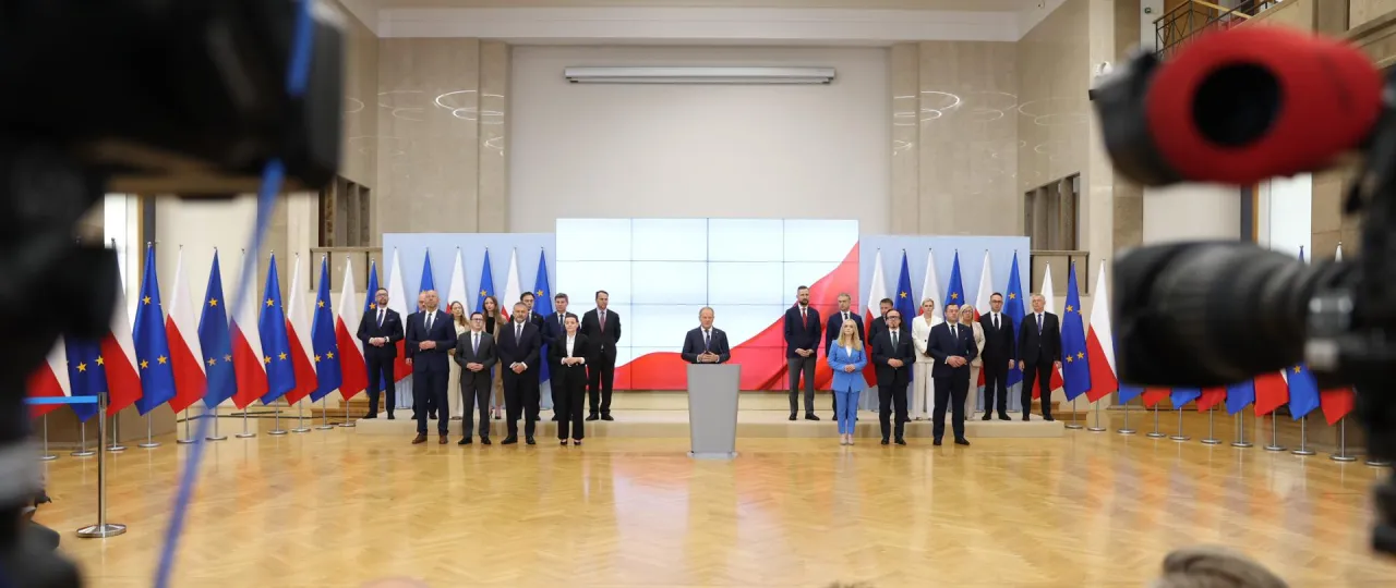 Grupa osób w garniturach i sukienkach stoi przed flagami Polski i UE. W tle ekran z biało-czerwoną flagą. Kto jest premierem Polski?