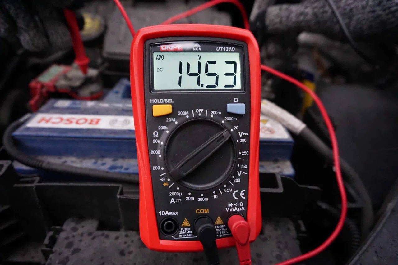 Multimetr pokazuje 14.53V, co jest kluczowe, jak sprawdzić alternator miernikiem. W tle widać akumulator samochodowy.