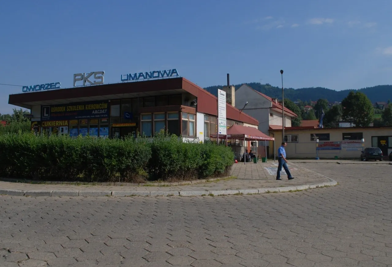 Limanowa dworzec autobusowy PKS. W tle budynki, drzewa i wzgórza. Na pierwszym planie brukowany plac.