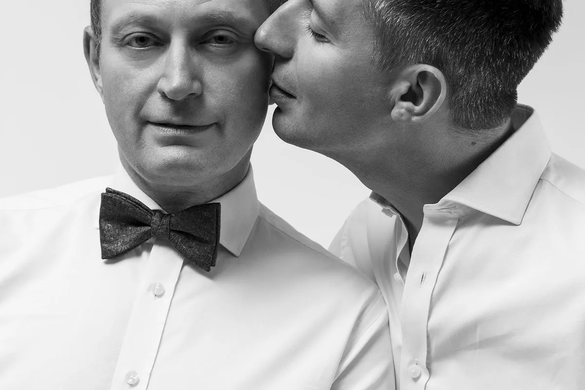 Czarno-białe zdjęcie dwóch mężczyzn w białych koszulach. Jeden z nich, z muszką, patrzy wprost, drugi całuje go w policzek. To Michał Cessanis żona.