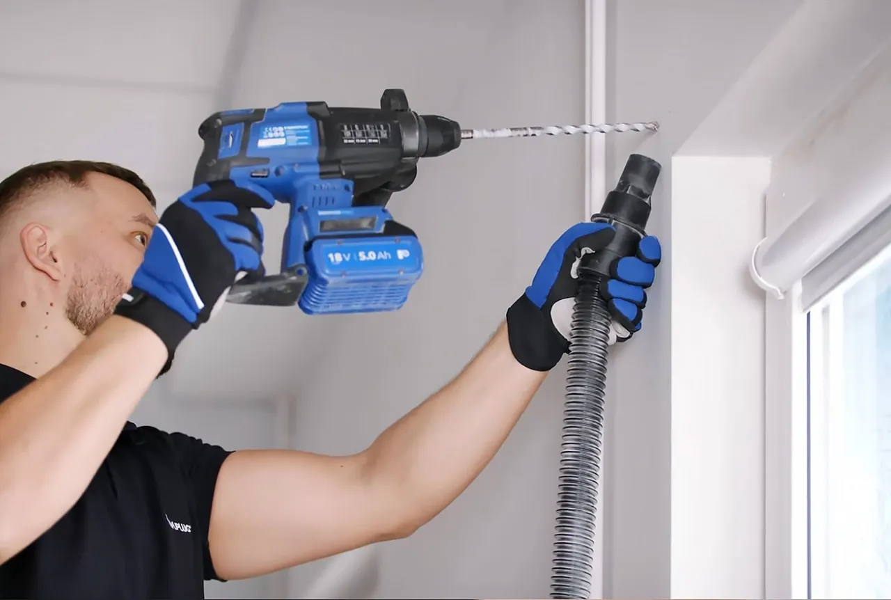 Mężczyzna w rękawicach wierci otw&oacute;r w suficie, przygotowując się do montażu karnisza sufitowego. Odkurzacz zbiera pył.