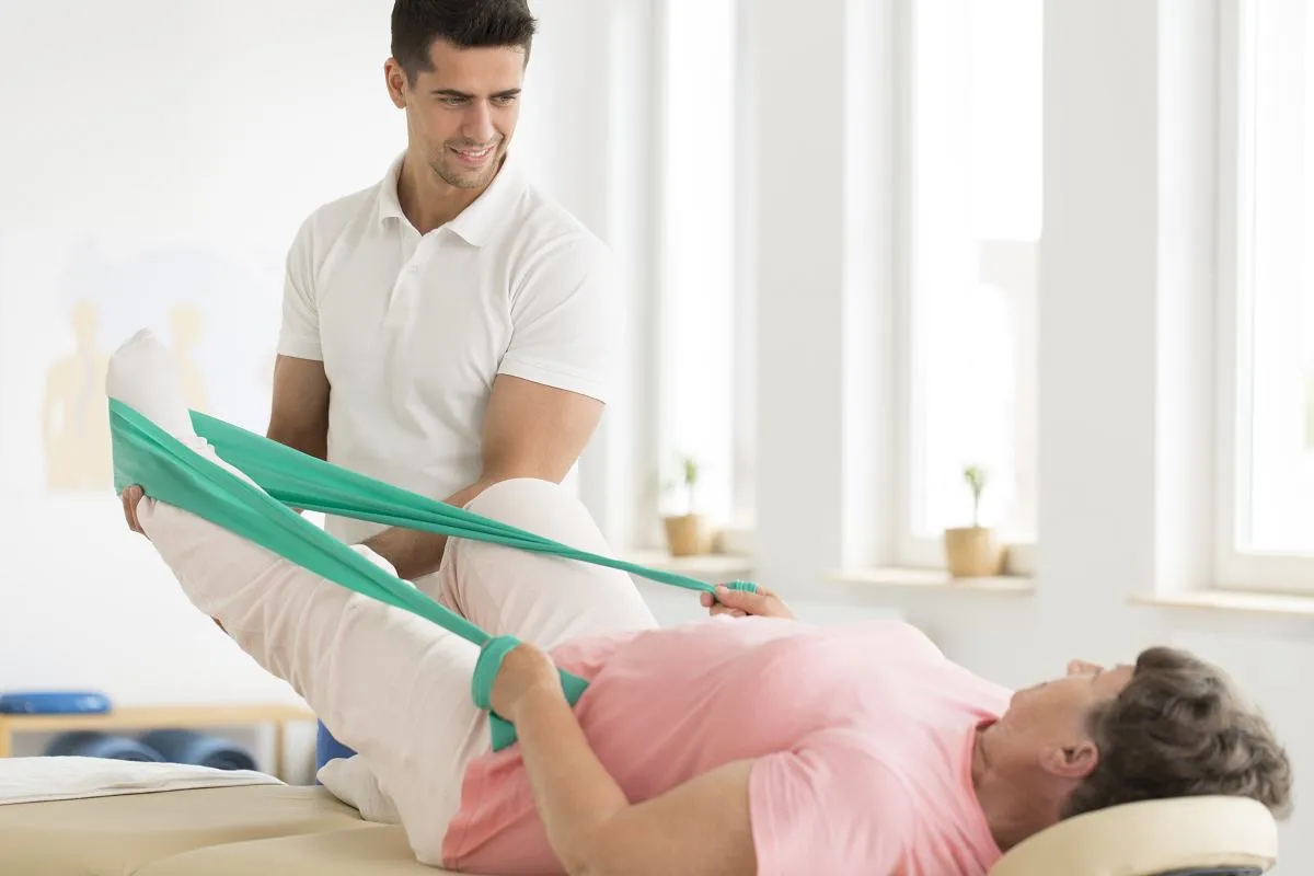 Fizjoterapeuta pomaga pacjentce w rehabilitacji po endoprotezie stawu biodrowego, ćwicząc nogę z użyciem taśmy.
