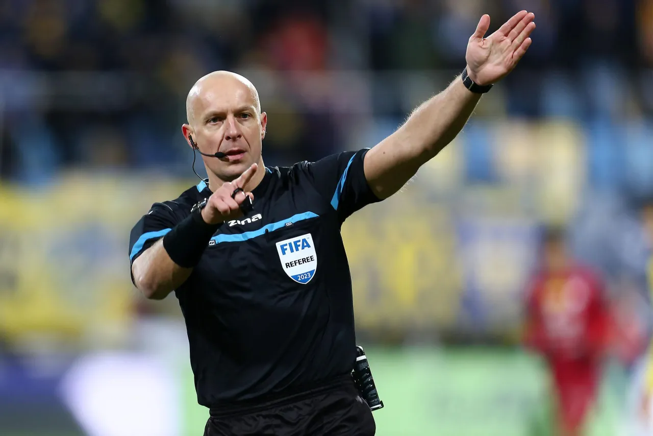 Sędzia z gwizdkiem i podniesioną ręką, w tle trybuny. Ciekawe, ile zarabia sędzia w ekstraklasie za takie emocje.